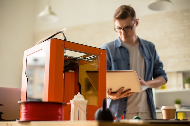 A person setting up a Creality Ender 3 V2 Neo 3D printer on a desk. The person is carefully connecting wires and adjusting the printer's bed. The background shows a room with various 3D printing tools and filament spools.