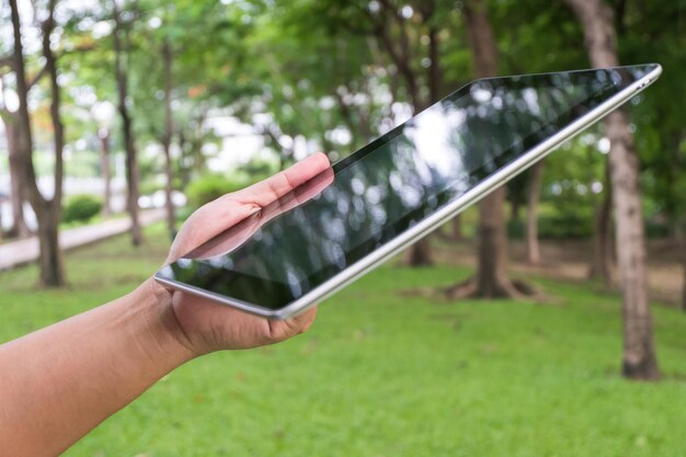 A close-up of a foldable portable solar panel charging a tablet outdoors, showcasing the panel's compact design and the tablet's charging indicator. The setting is a park with trees in the background.