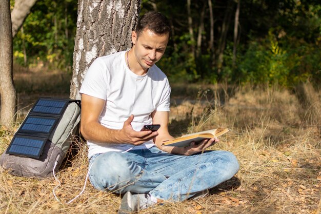 A person sitting at a campsite with a portable solar panel array charging multiple devices (smartphone, power bank, and camping lantern). The scene is set at dusk, highlighting the importance of off-grid power solutions.