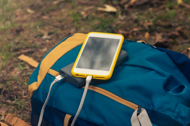 A close-up of a portable solar charger attached to a backpack, charging a smartphone. The background shows a sunny hiking trail.