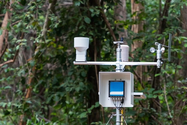 A close-up view of an automated weather station in a rural setting. The station includes various instruments such as anemometers, thermometers, and barometers. The background shows a clear blue sky and green fields.